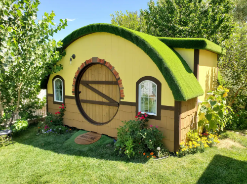 A whimsical yellow hobbit-style house with a grassy roof, surrounded by colorful flowers and greenery.